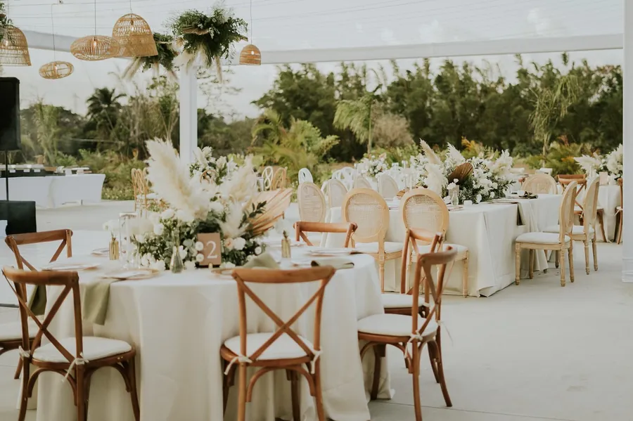 Mesas redondas con manteles blancos, sillas de madera y arreglos de flores bajo la carpa