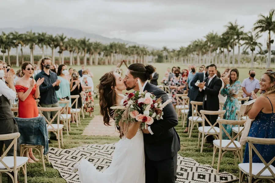 Novios besándose al salir de la ceremonia, entre invitados y con la fila de palmeras detrás