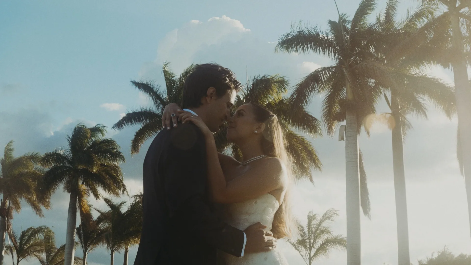 Novios abrazados bajo las palmeras altas, con el cielo de la tarde